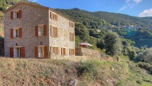 Un bâtiment en pierre doté de nombreuses fenêtres et volets, entouré d’un paysage vallonné.