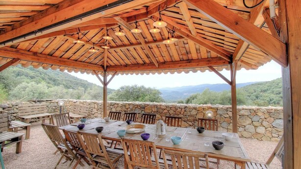 Une pergola en bois installée à l’extérieur, avec une table dressée pour les repas, des murs en pierre et une vue panoramique.