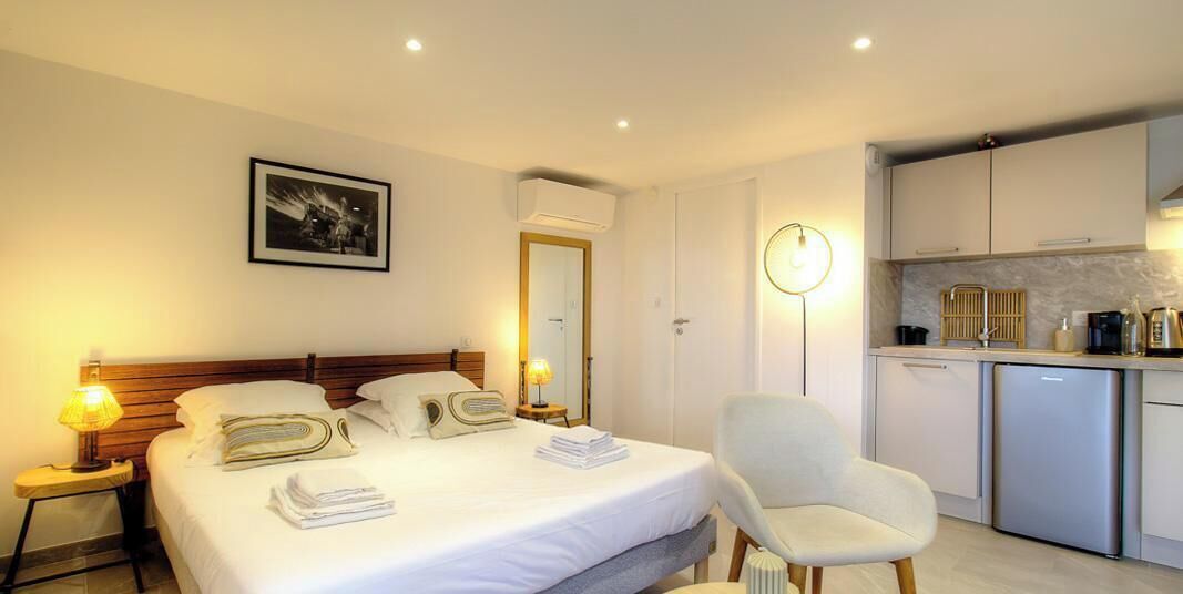 Une chambre d’hôtel comprenant un lit, des lampes de chevet, une chaise, un bureau, un coin cuisine avec un réfrigérateur et une horloge murale.