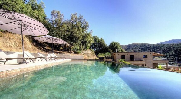 Una piscina infinita cristalina con sillones de descanso y sombrillas, rodeada por una colina y un edificio de techo plano.