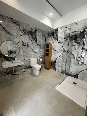 A modern bathroom with a marble wall, a round mirror, a toilet, a sink, and a glass-enclosed shower.