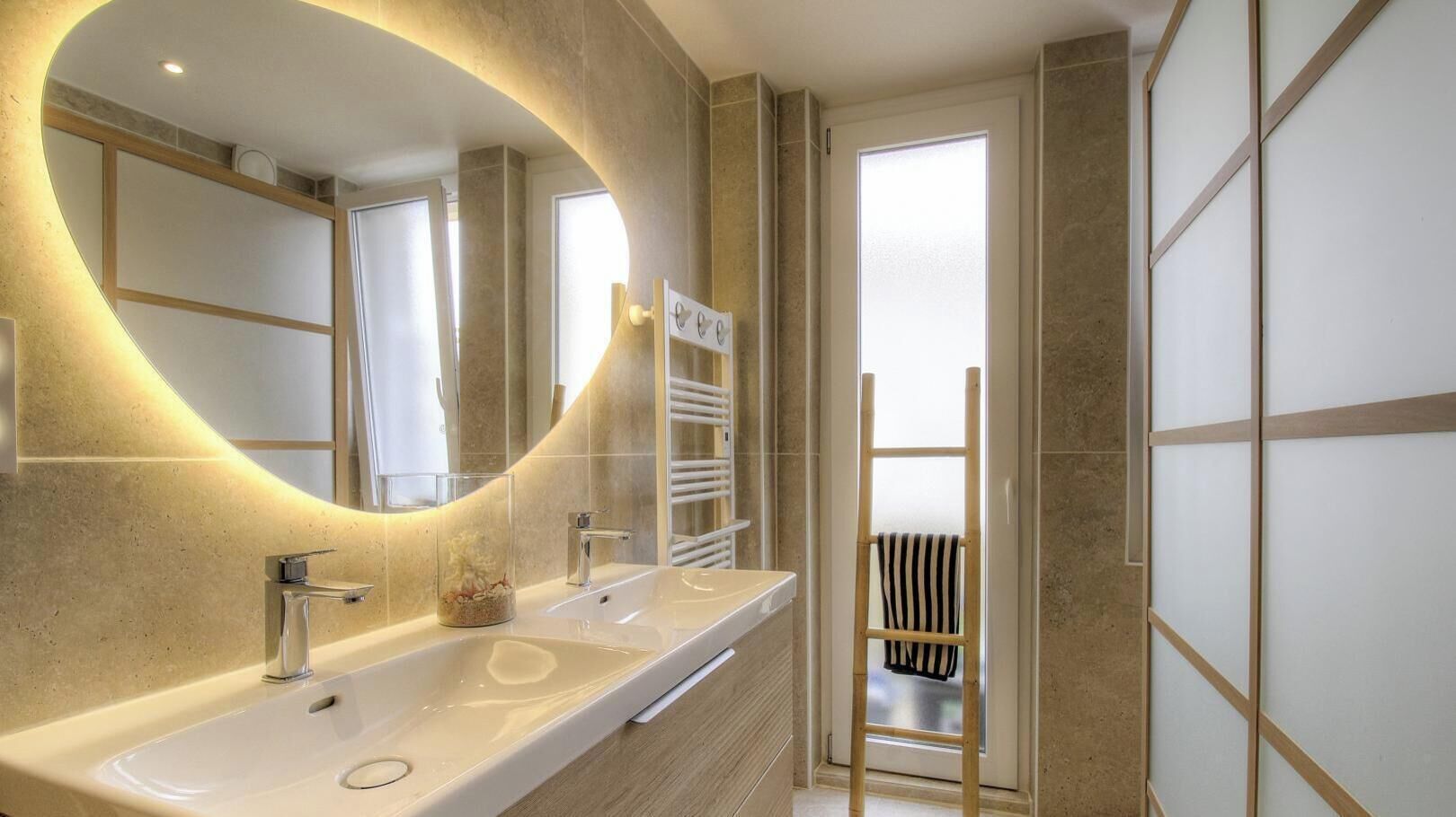 A modern bathroom with a large oval mirror, two sinks, and a towel rack.