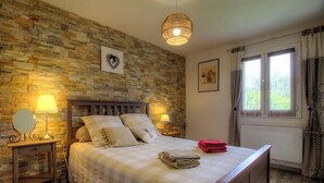 Un dormitorio con pared de piedra, una cama con almohadas, una mesita de noche con lámpara y una ventana con cortinas.