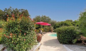 Eine Holzterrasse führt zu einem Pool mit einem roten Sonnenschirm, umgeben von üppiger Vegetation.