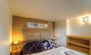 Ein Schlafzimmer mit einem Bett, einem Kopfteil, einem Nachttisch, einer Lampe und einem an der Wand montierten Regal.