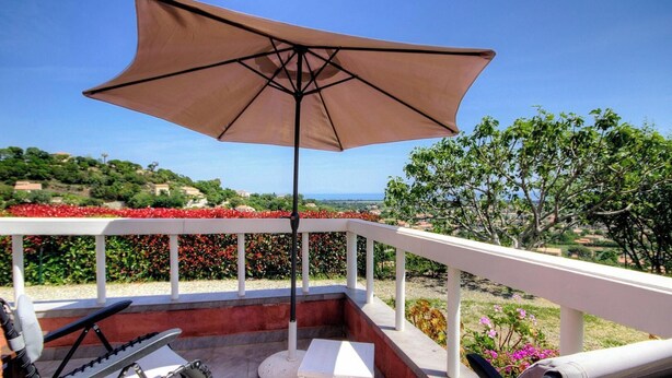 Ein Balkon mit einem beigen Sonnenschirm, weißem Geländer und Blick auf einen angelegten Garten sowie entfernte Hügel.