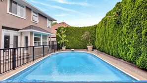 Une piscine entourée d’une clôture en métal noir, une maison à l’extérieur beige et une haie bien entretenue.
