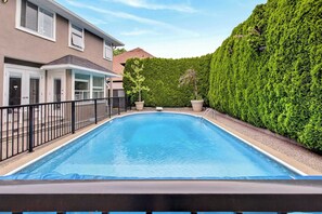 A swimming pool with a black metal fence, a house with a beige exterior, and a well-maintained hedge. - 2BR Surrey Pool Suite Near White Rock (Surrey)