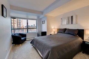 Un dormitorio con una cama grande, una silla, una mesita de noche, una lámpara y una ventana con vistas a la ciudad.