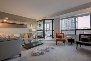 Una sala de estar moderna con sofá, mesa de centro y sillas, un separador de vidrio y ventanas grandes con vistas a la ciudad.