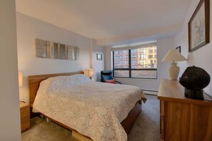A hotel room with a bed, a desk with a lamp, a chair, and a window with a city view.