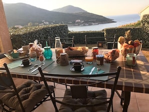 Ein Frühstückstisch mit verschiedenen Speisen und Getränken, mit Blick auf eine Küstenlandschaft und Berge.