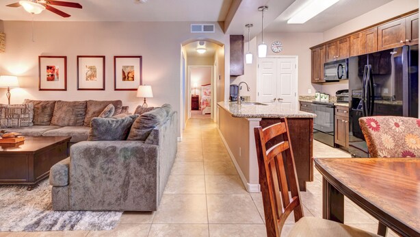 Una sala de estar moderna con un sofá, una mesa de centro y una zona de comedor con una mesa de madera. Se ve una cocina con refrigerador y microondas al fondo.