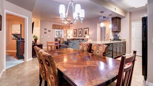 Un comedor amplio con una gran mesa de madera, rodeado de sillas con diseños, una lámpara de araña y una cocina al fondo.