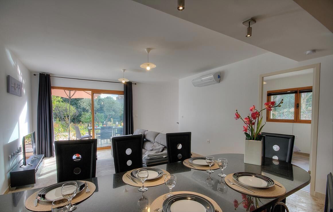 A dining area with a round table set for four, a sofa in the background, and a view of greenery outside.