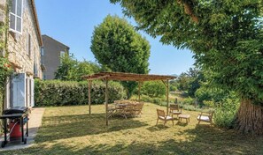 Un espace extérieur abrité, avec une table et des chaises, entouré de verdure et d’un bâtiment.