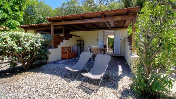 Ett takförsett utomhusområde med sittplatser, bestående av två solstolar, en träpergola och en trappa som leder till en dörr.