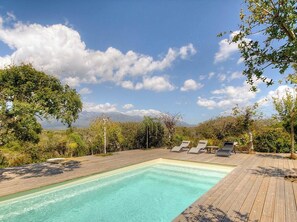 Ein rechteckiger Pool mit Holzterrasse, umgeben von Grünflächen und Bäumen, mit blauem Himmel darüber.