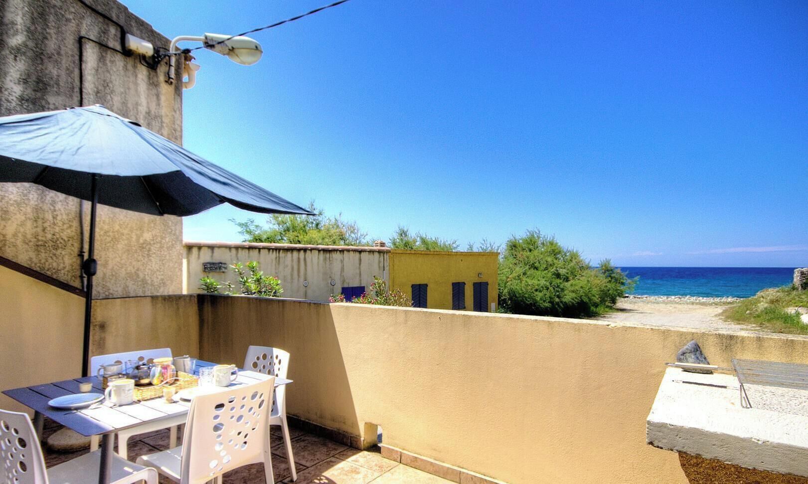 A patio with a table and chairs, a blue umbrella, and a view of the sea.