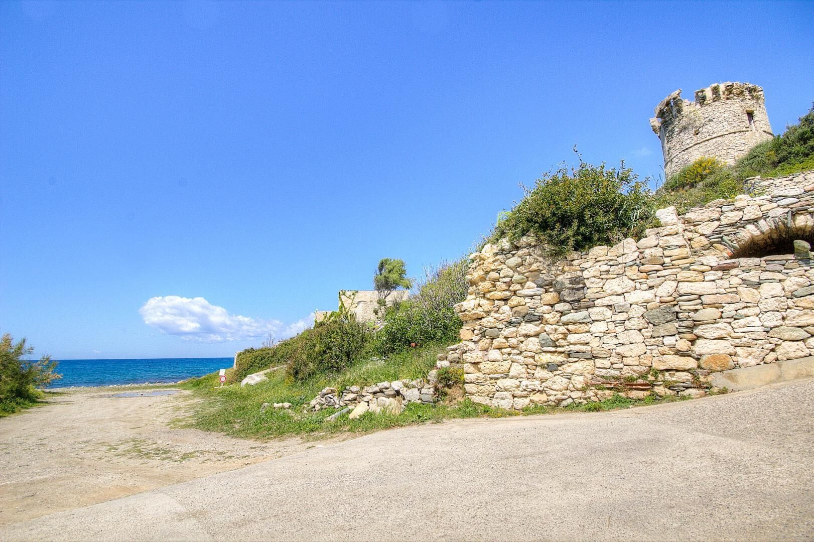 Eine Küstenstraße mit einer Steinmauer und einem Turm, die zu einem Strand führt.