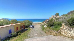 A coastal road leading to a beach with a car parked on the side, a small building with red shutters, and a stone wall. - Mare e Torre - FARINOLE (FARINOLE)