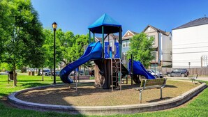 Une aire de jeux avec des toboggans et une structure de jeux couverte, entourée de pelouse et d’arbres.