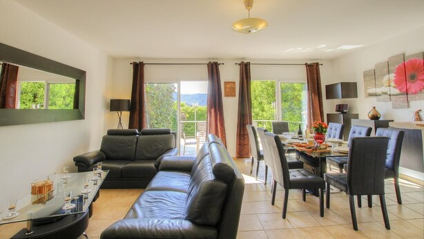 En moderne stue med sofa, spisebord, stole og et stort vindue med gardiner.