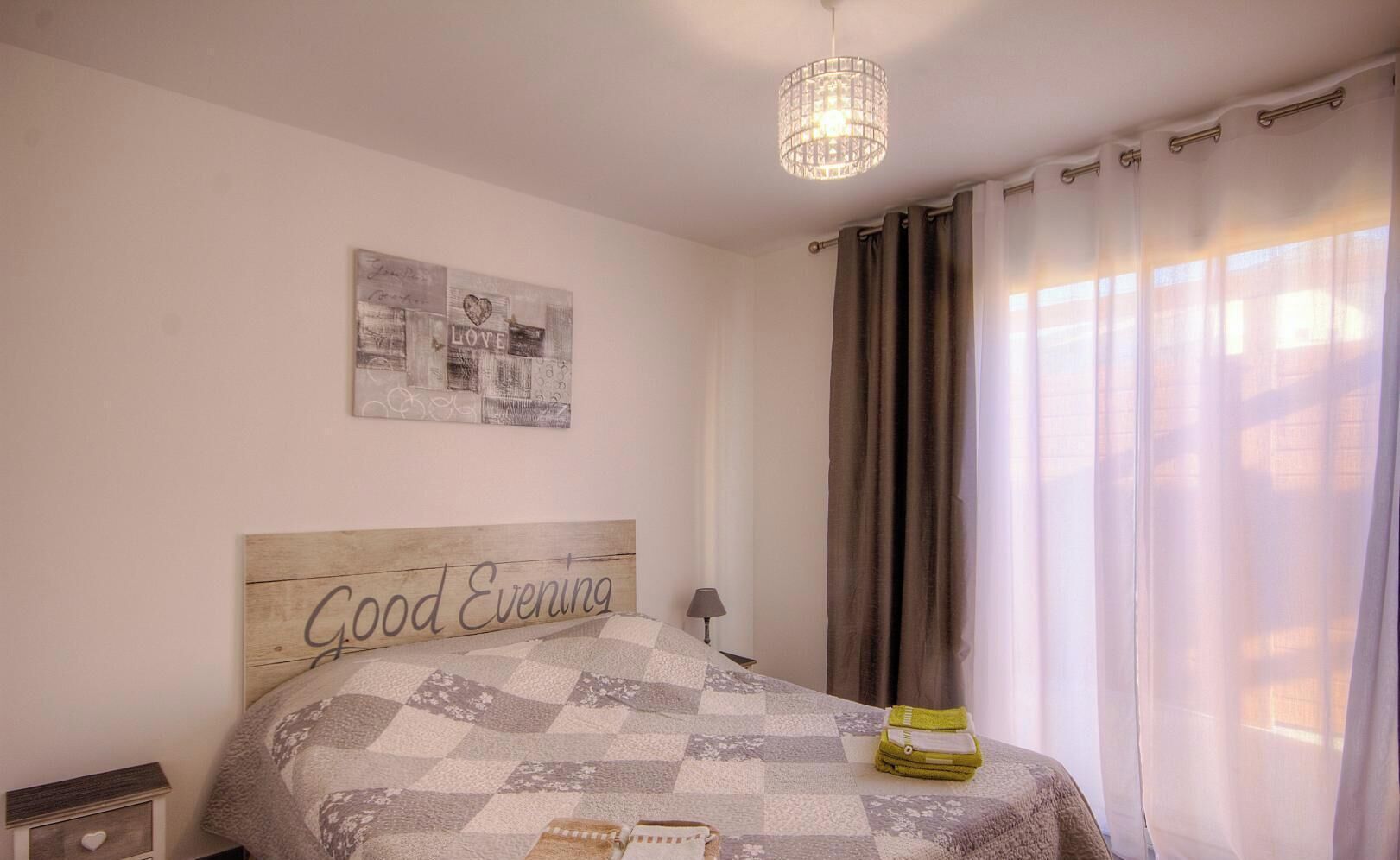A bedroom with a bed, a headboard with 'Good Evening' text, a wall art piece, a lamp, and curtains.