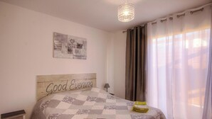 Quarto com cama, cabeceira com o texto 'Good Evening', uma obra de arte na parede, abajur e cortinas.