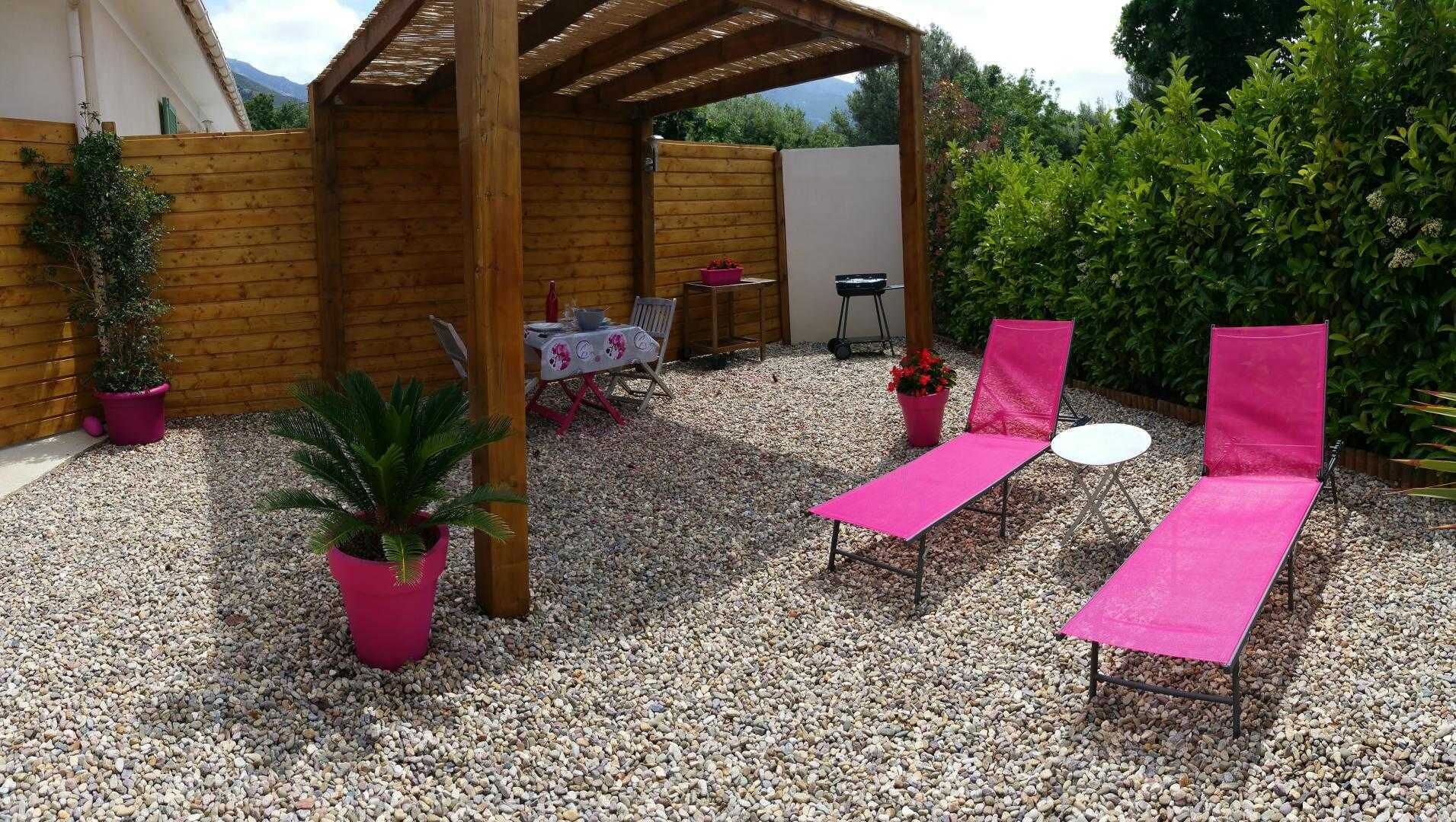 Eine überdachte Terrasse mit Tisch und Stühlen, umgeben von Topfpflanzen und einem Kiesgrund.