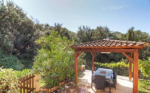 Eine hölzerne Pergola mit Ziegeldach, einem Tisch mit Stühlen und einem Garten mit verschiedenen Pflanzen.