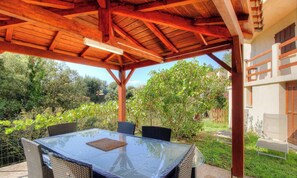 Eine überdachte Terrasse im Freien mit einem Glastisch und Korbstühlen, umgeben von Grünpflanzen und einem Gebäude mit Fenstern.