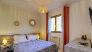 Ein Schlafzimmer mit einem Bett, Nachttischlampen, einem runden Spiegel, einem Fenster mit Vorhängen und einem kleinen Tisch mit einem dekorativen Gegenstand.