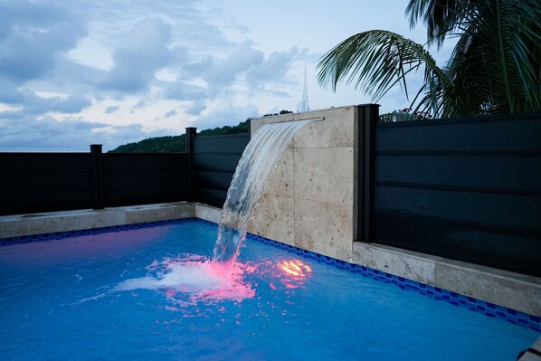 A swimming pool with a waterfall feature, surrounded by a low wall and palm trees. - Villa with Private Pool and Sea View (Luquillo)