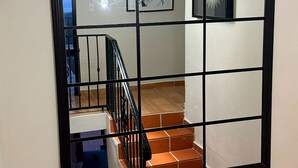 Un grand miroir à cadre noir reflète un escalier, un luminaire et un tableau accroché au mur.
