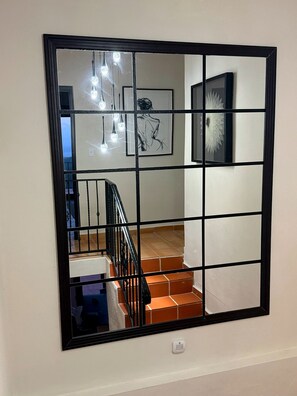 Un espejo grande con marco negro que refleja una escalera, una lámpara y un cuadro colgado en la pared.