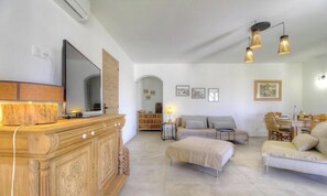 Ein modernes Wohnzimmer mit Flachbildfernseher, Holzschrank, Sofa, Hocker, Esstisch und Wandleuchten.