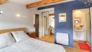 A bedroom with a bed, wooden beams, a bathroom with a sink and mirror, and a door leading to another room. - Nanu - AULLENE - Gîte (AULLENE)