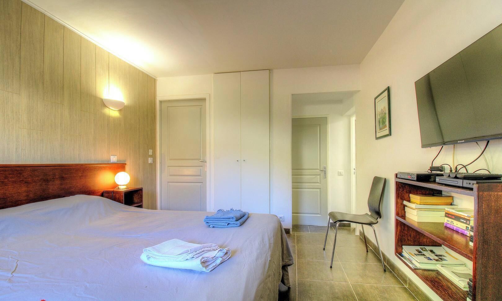 A hotel room with a bed, a desk with a chair, a bookshelf, and a television.