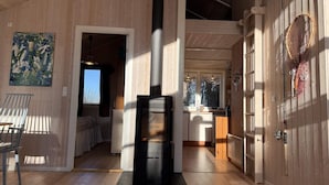 A wooden interior with a central fireplace, a dining area with a chair, and a kitchen with a window. - 4 Star Holiday Home in Kalundborg (Kalundborg)