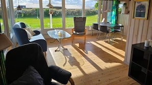 A spacious room with a large window, a sofa, a glass coffee table, a dining table with chairs, and a hanging light fixture. - 4 Star Holiday Home in Kalundborg (Kalundborg)