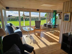 Ein geräumiges Zimmer mit großem Fenster, einer Couch, einem Glastisch, einem Esstisch mit Stühlen und einer Pendelleuchte.
