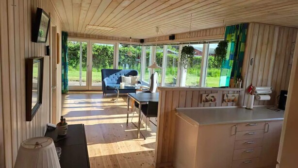 A room with a wooden ceiling, a sofa, a dining table, a kitchen area, and large windows offering a view of greenery. - 4 Star Holiday Home in Kalundborg (Kalundborg)