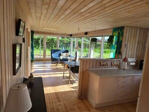 A room with a wooden ceiling, a sofa, a dining table, a kitchen area, and large windows offering a view of greenery. - 4 Star Holiday Home in Kalundborg (Kalundborg)