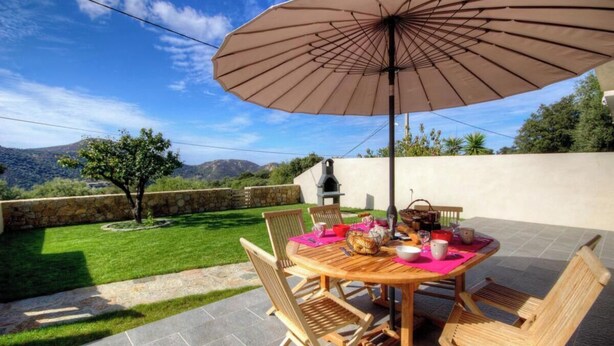 Um pátio externo com uma mesa de madeira redonda, cadeiras, um grande guarda-sol e um muro de pedra.