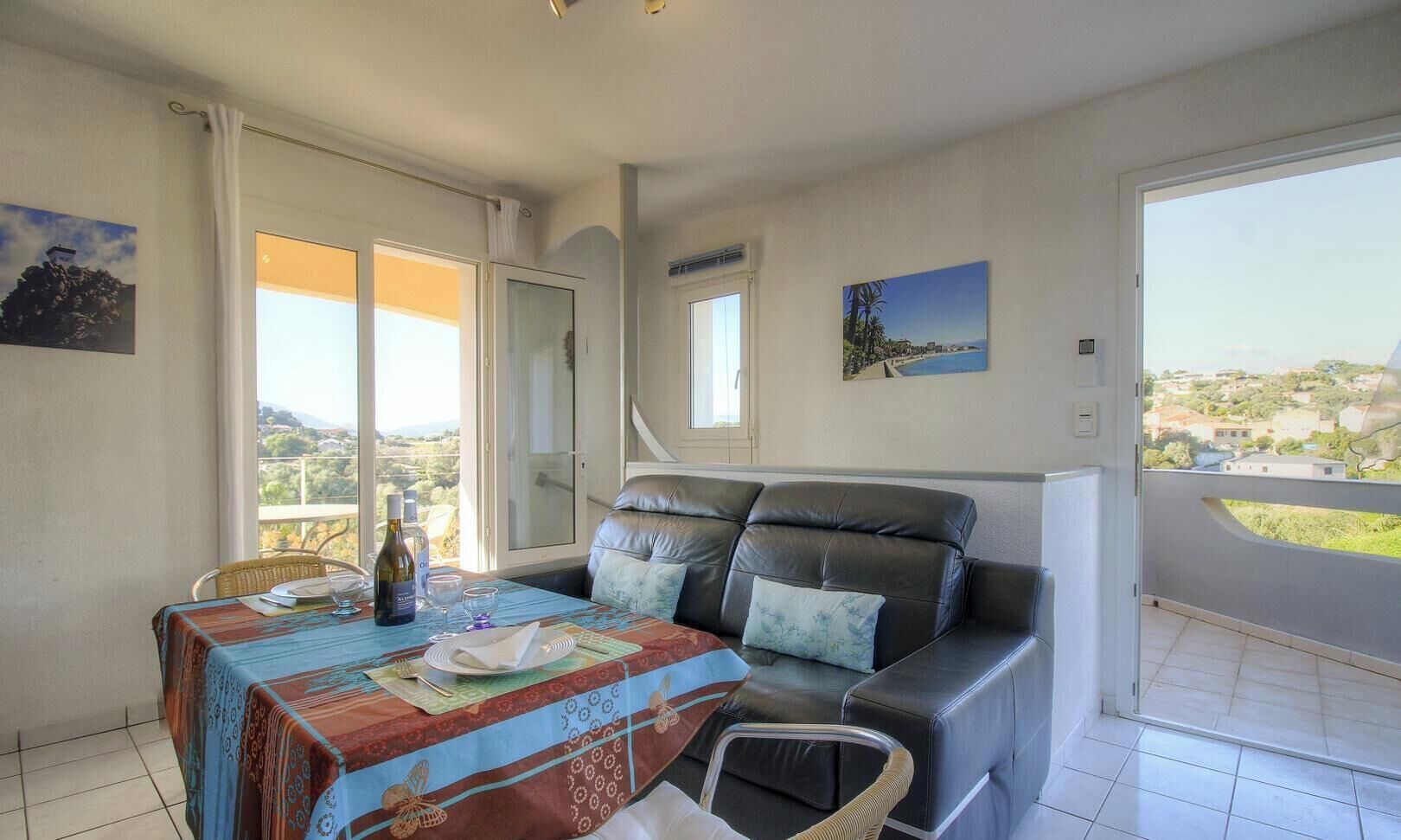 A balcony with a view of a landscape, a dining table with a tablecloth, a sofa with cushions, and a chair.
