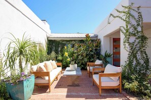 Eine Terrasse im Freien mit einer Couch, Stühlen und einem Tisch, umgeben von Topfpflanzen und einer weißen Wand.