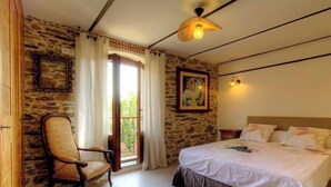 Quarto com parede de pedra, cama com roupa de cama branca, cadeira de madeira, pintura e janela com cortinas.