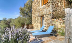 Ein Steinhaus mit einem hölzernen Tor, zwei blauen Liegestühlen und violetten Blumen.