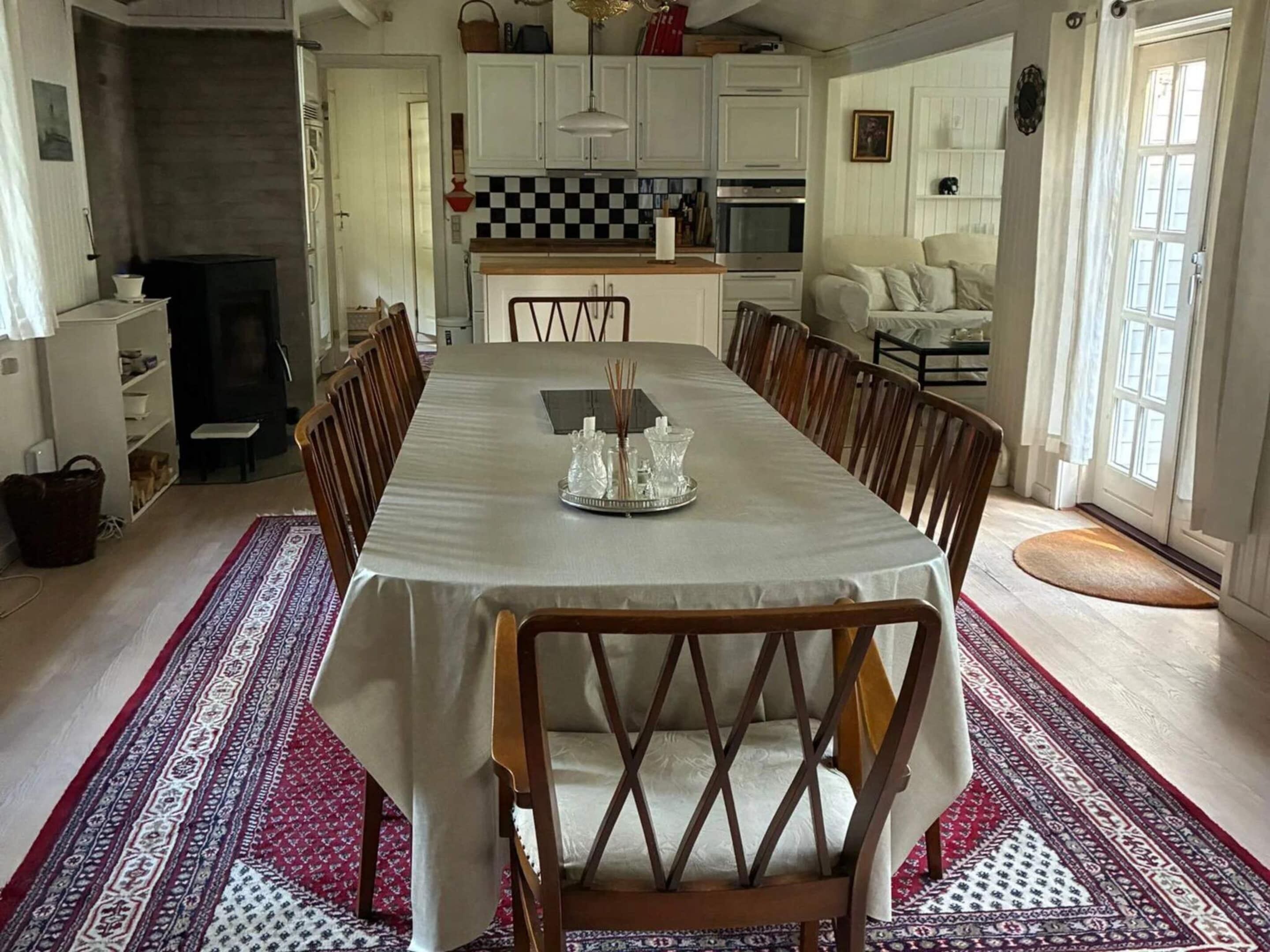 A dining room with a long table, chairs, a kitchen in the background, and a patterned rug.